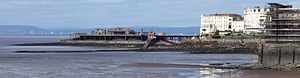 Structure supported on metal legs coming out of the sea, stretching from the land showing several white buildings to an island with buildings on it. A lifeboat house with slipway is attached to the side. In the distance across the water are hills and buildings.