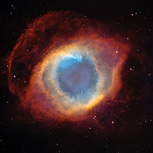 Colorful shell which has an almost eye like appearance. The center shows the small central star with a blue circular area that could represent the iris. This is surrounded by an iris like area of concentric orange bands. This is surrounded by an eyelid shaped red area before the edge where plain space is shown. Background stars dot the whole image.