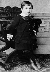 A young boy with short hair and a round face, wearing a white collar and large bow, with vest, coat, skirt and high boots. He is leaning against an ornate chair.