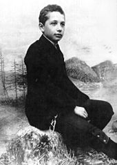 Studio photo of a boy seated in a relaxed posture and wearing a suit, posed in front of a backdrop of scenery.