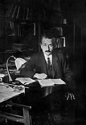 Einstein, sitting at a table, looks up from the papers he is reading and into the camera.