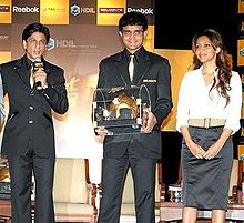 Two middle-aged males and one female standing. The man in the middle wears a black suit and carries a golden colored casket. The other man to his right wears a black suit and speaks in a microphone. The lady on the left wears a white shirt and black skirt. Her hair is brownish and falls in locks around her.