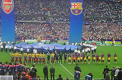 Two teams of sportsmen, one in yellow shirts and one in red and blue shirts, stand in a line. In front of the line is a line of children in equivalent shirts, but each in front of the opposite team.  A number of other people stand in the foreground looking on. Behind the teams, a number of people hold a large circular piece of fabric with logos printed on, and two large banners hang down vertically from above the whole scene.