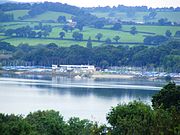 Low concrete building on the edge of the water, with boats on either side. Vegetation in the foreground, hills in the background